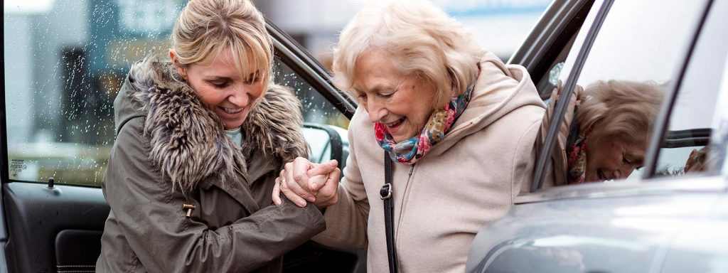 Young woman helps senior woman out of car.