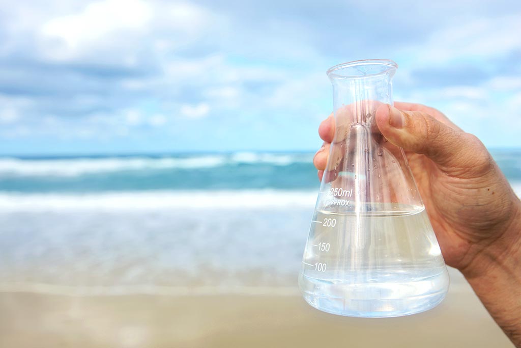 A hand holding a beaker of water with a beach in the background.