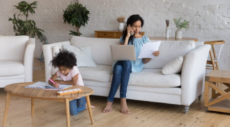 Mother focused on work with phone and papers in hand, and laptop on her lap while child is doing homework on the coffee table