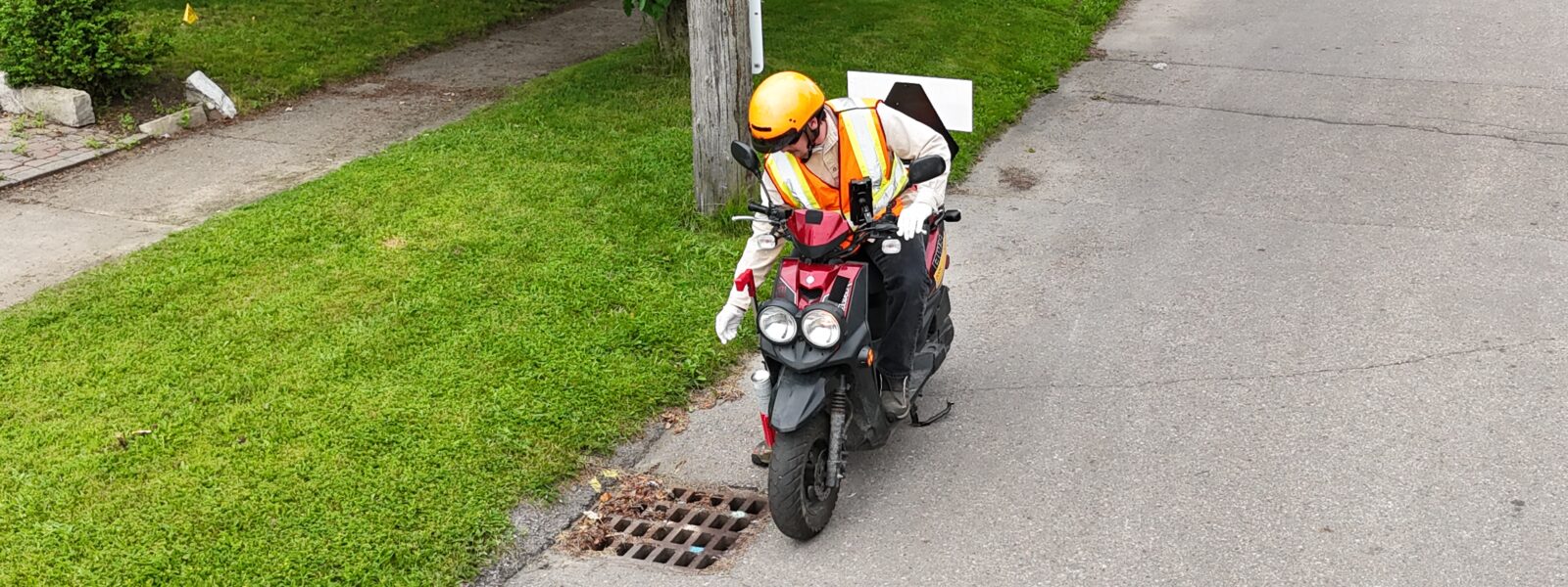 Man with helmet and safety vest sprinkling larvicide to roadside catch basin, while riding a motorized scooter.