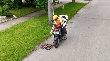 Man with helmet and safety vest sprinkling larvicide to roadside catch basin, while riding a motorized scooter.