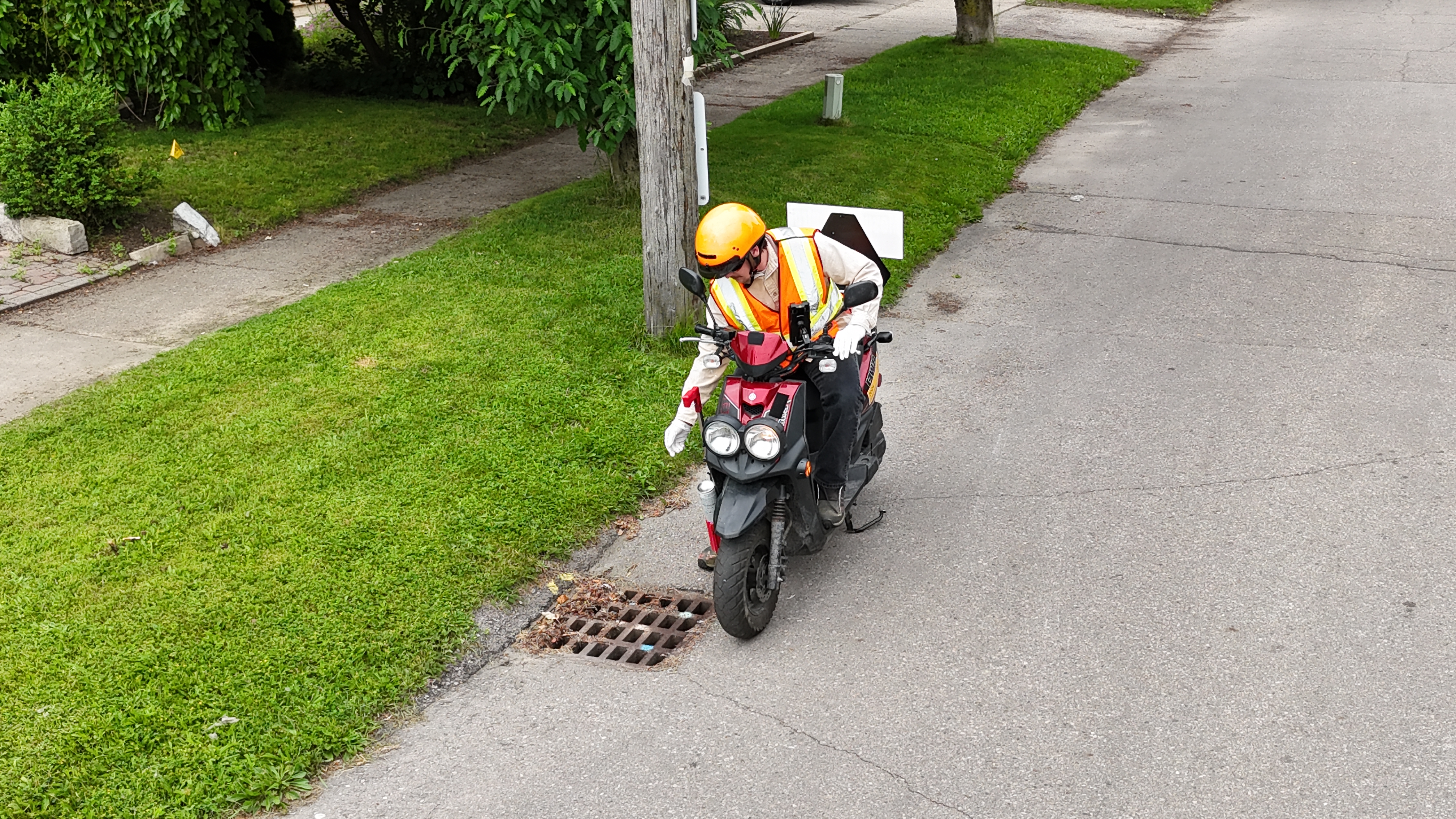 Man with helmet and safety vest sprinkling larvicide to roadside catch basin, while riding a motorized scooter.